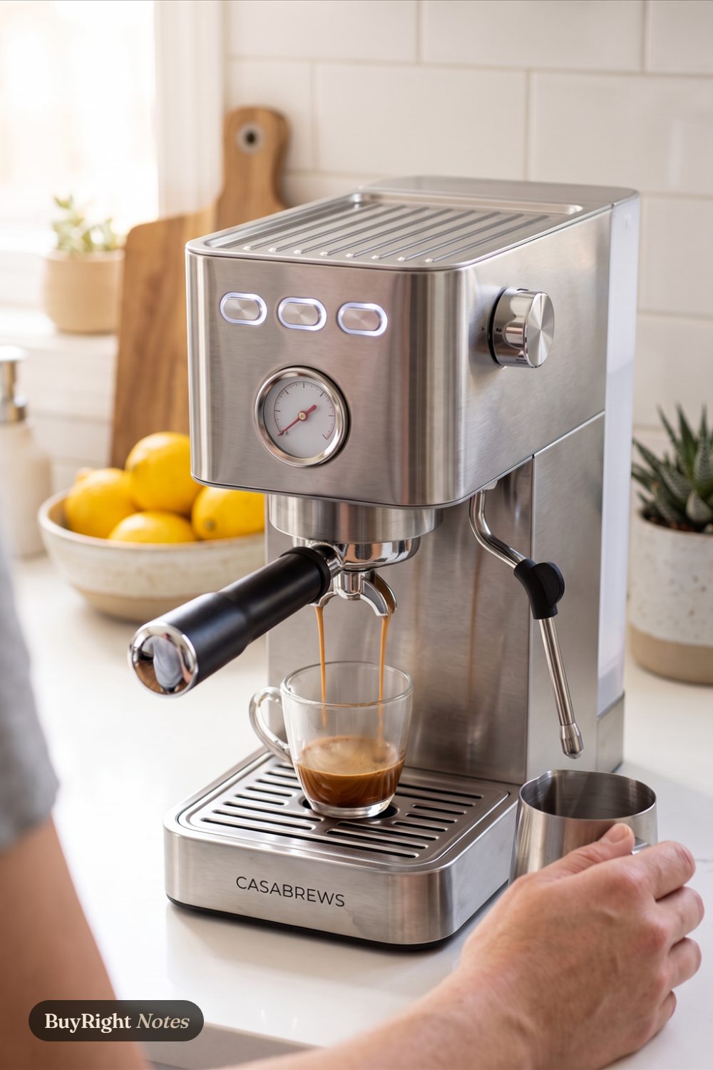 Espresso pours in two streams from a CASABREWS CM5418 espresso machine into a glass cup while a hand holds a milk pitcher on a white apartment counter with lemons behind.