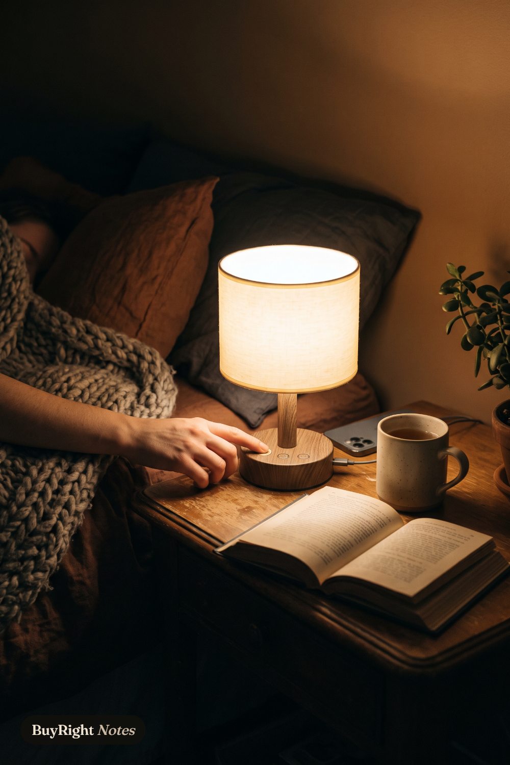 A hand reaches from under a chunky knit blanket to touch the base of a small glowing Fenmzee bedside lamp, an open book, a mug of tea and a phone charging on the nightstand.