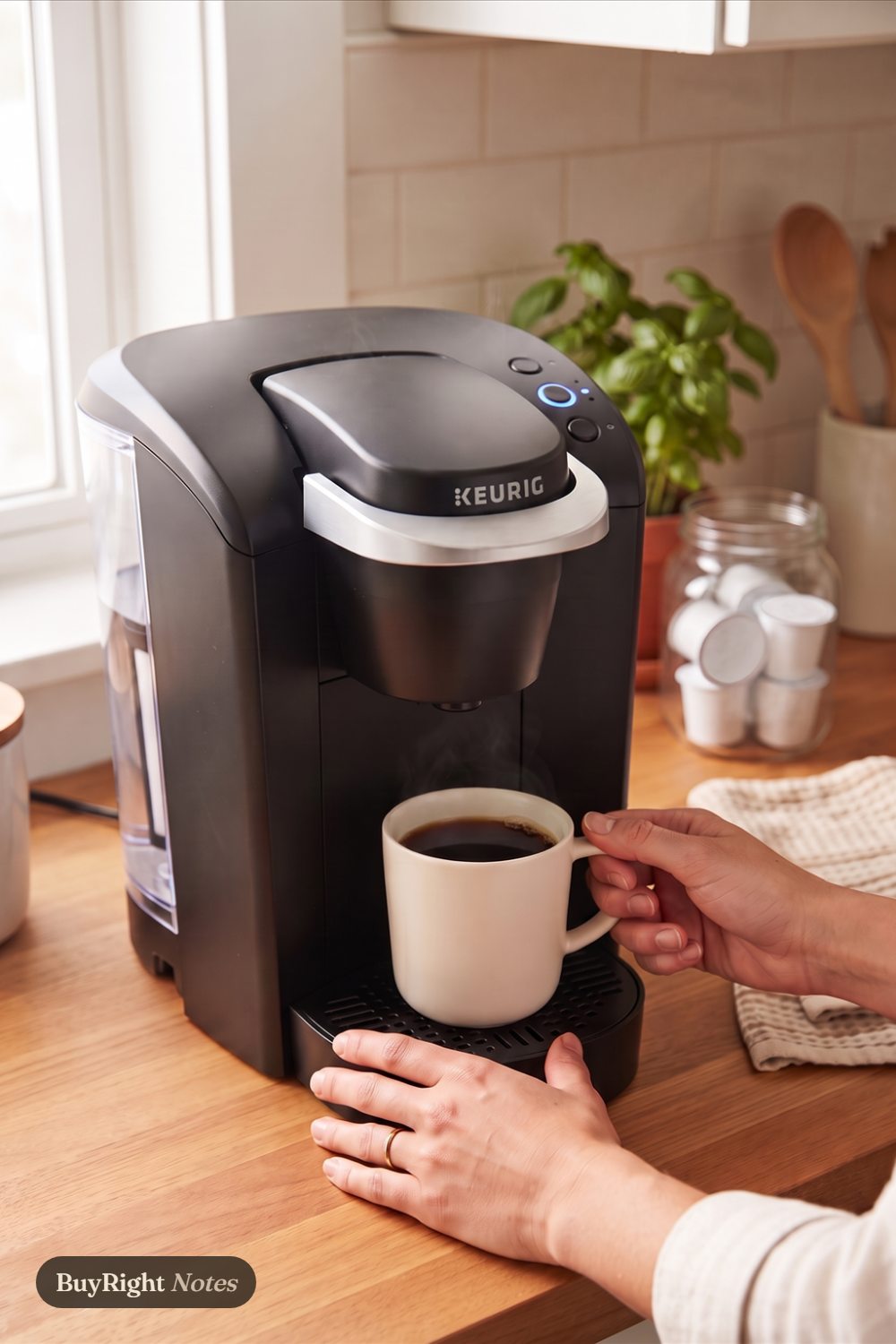 Hands lifting a cream mug of fresh coffee from a black Keurig K-Classic on a wood counter by a sunny window, with basil and a jar of plain pods beside it.