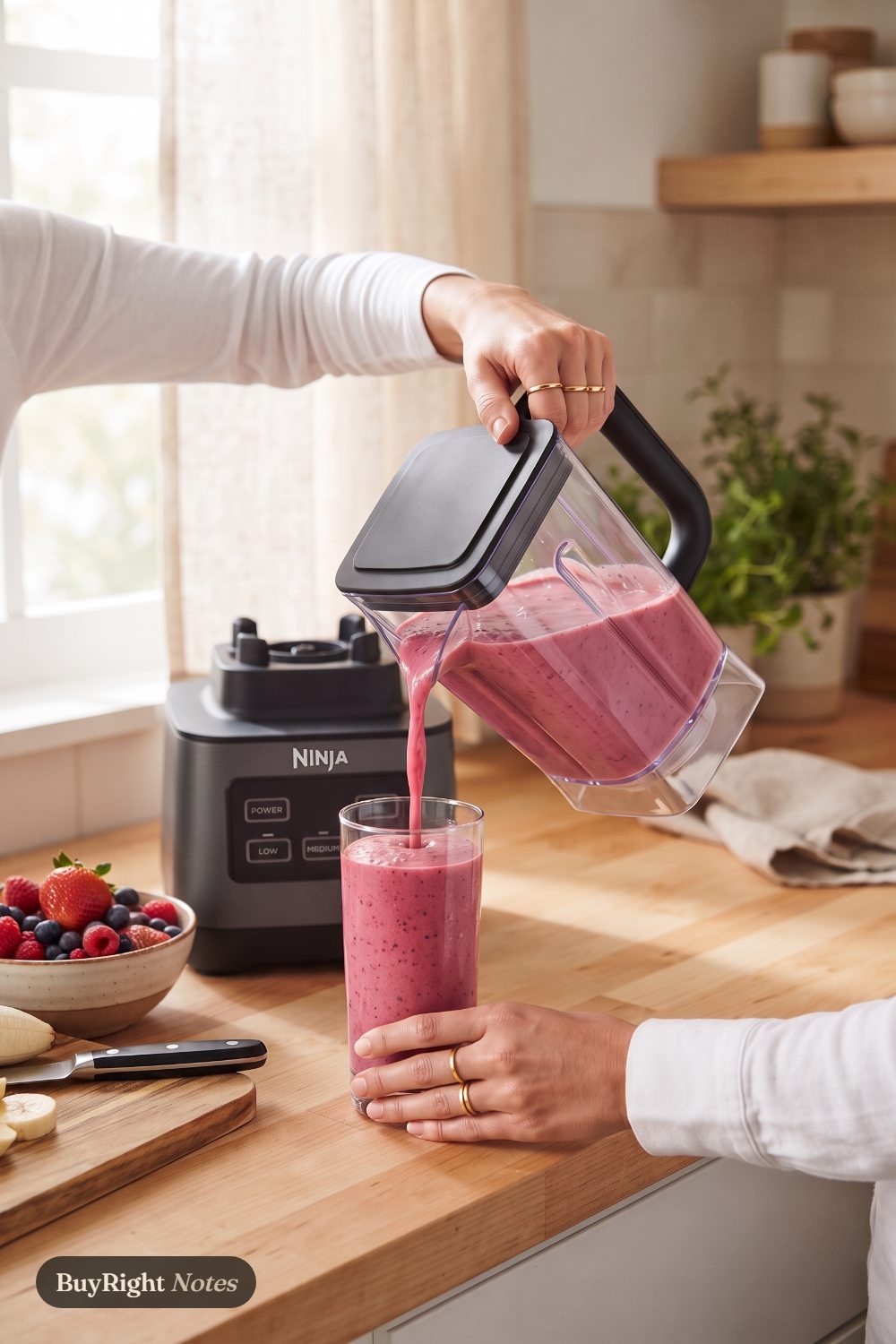 Hands pour a pink berry smoothie from a Ninja Professional blender pitcher into a tall glass on a bright kitchen counter, the Ninja base behind.