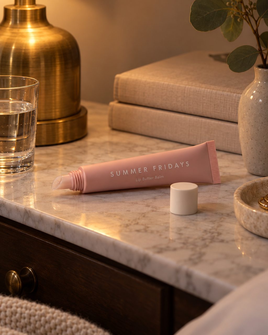 A Summer Fridays Lip Butter Balm tube on a bedside table at night beside a lamp, a glass of water and a folded blanket.